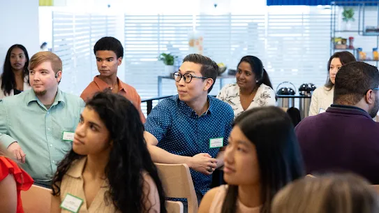 A group of various adults, possibly students or attendees, are seated in a room, with some looking forward and others looking to the side.